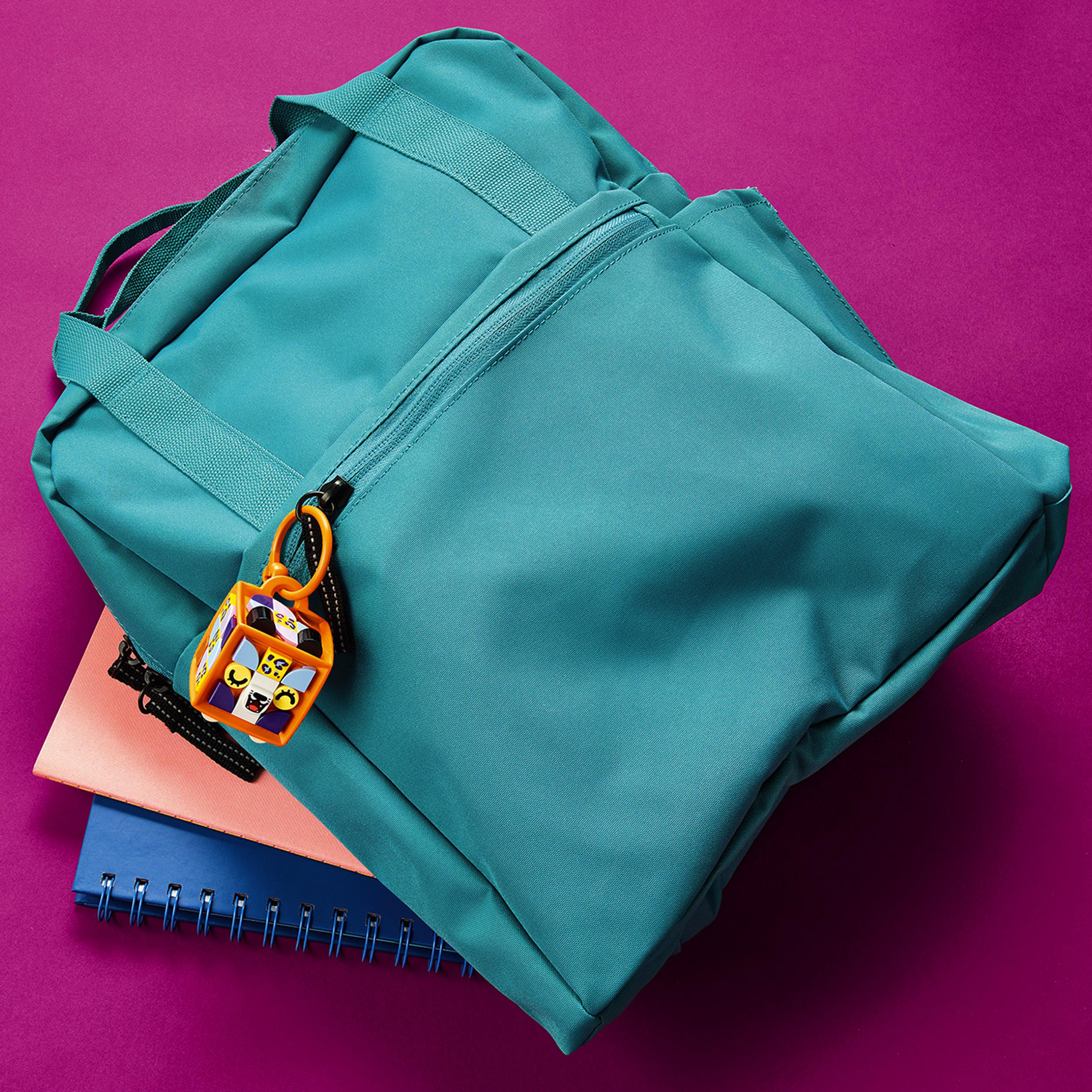 Teal backpack with a LEGO® DOTS Leopard bag tag, resting on colorful notebooks against a vibrant pink background.