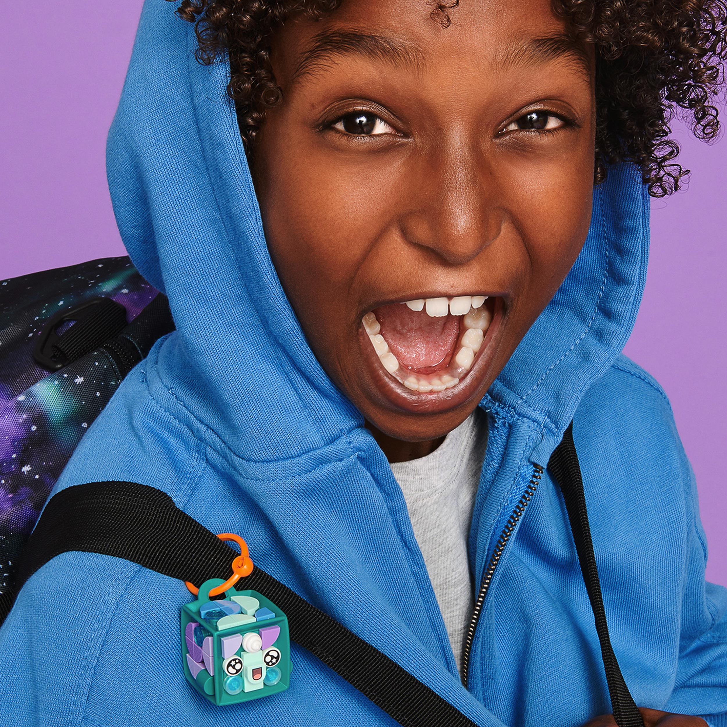 Child wearing blue hoodie with LEGO DOTS Narwhal bag tag attached to backpack, smiling excitedly.