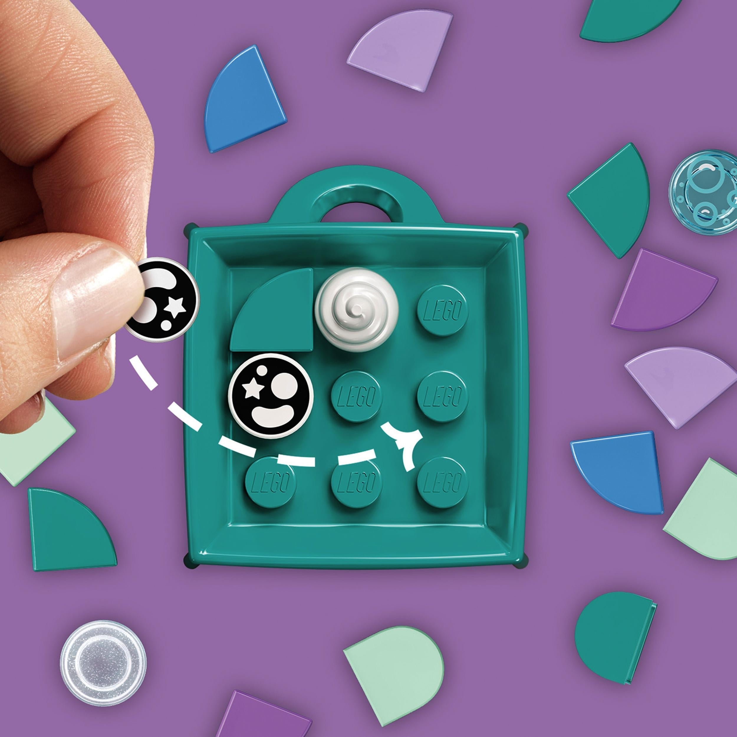 Child's hand customizing LEGO DOTS Narwhal Bag Tag craft kit with colorful tiles and tray.