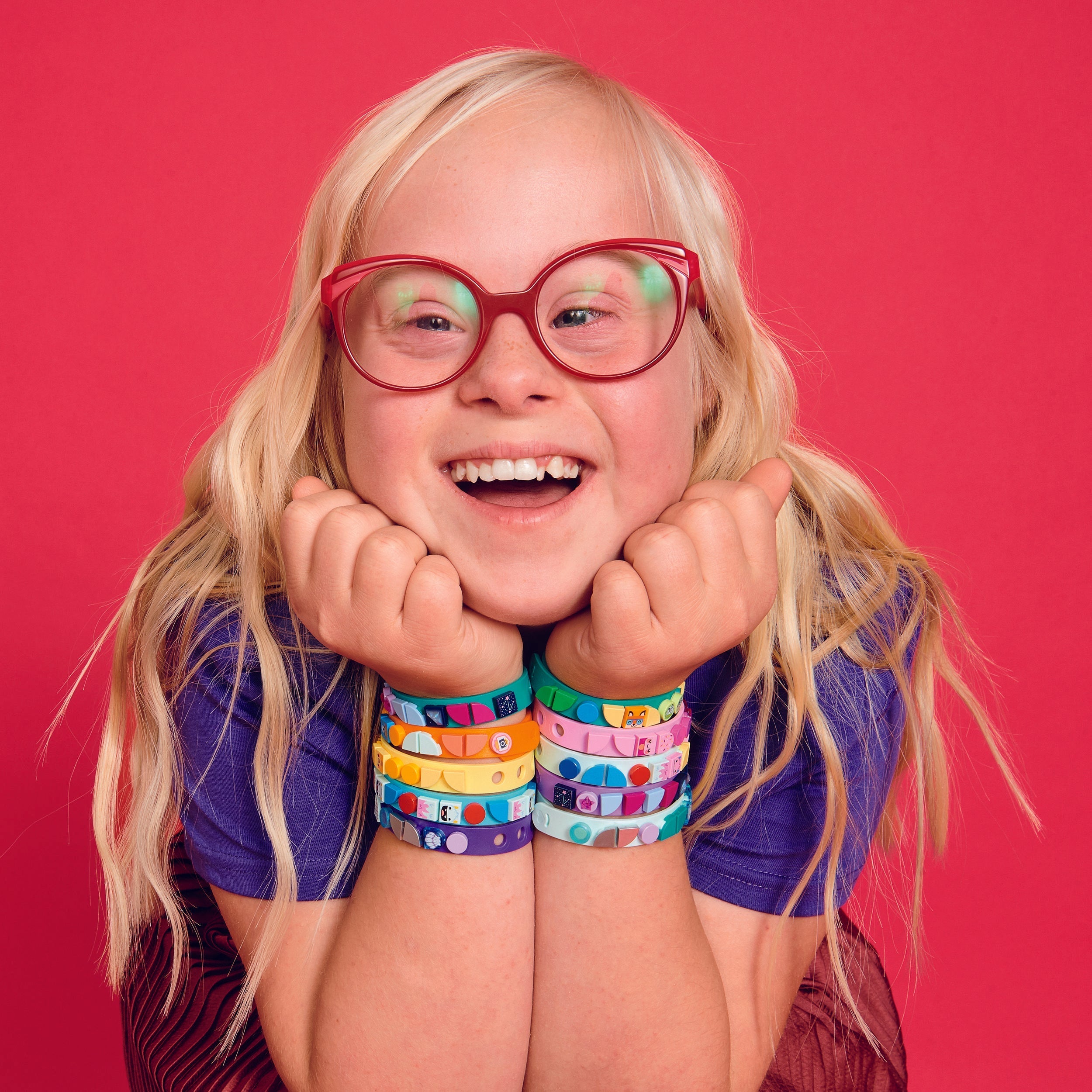 Child with colorful LEGO® DOTS starlight bracelets, smiling joyfully against a vibrant pink background.