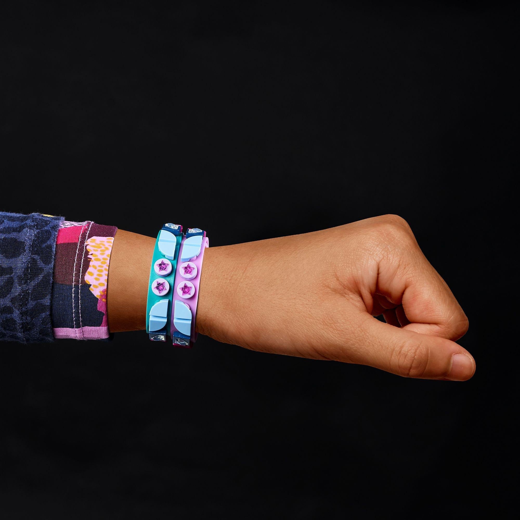 Child’s hand wearing LEGO® DOTS Starlight Bracelets in teal and purple with star tiles against a black background.