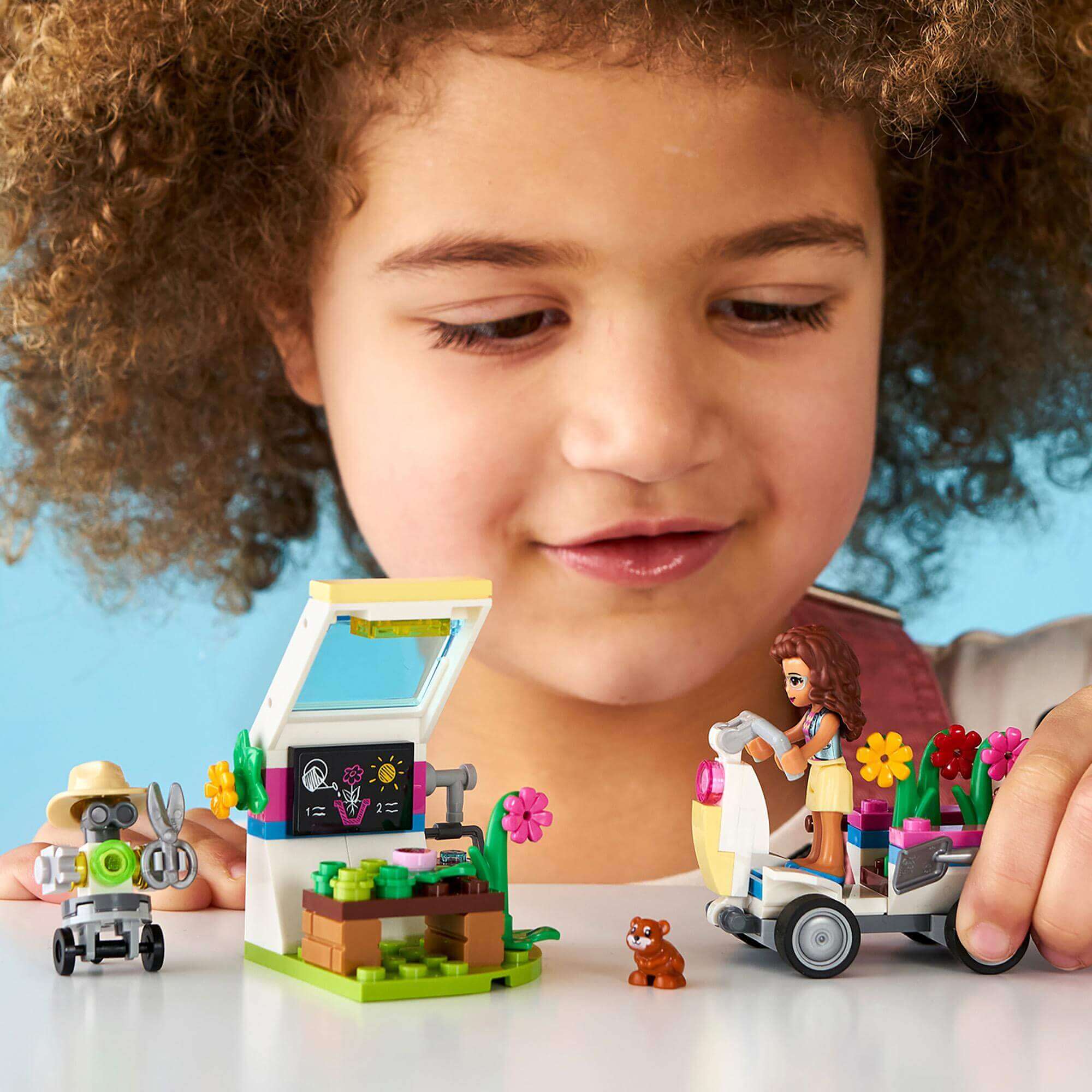Child playing with LEGO® Friends Olivia's Flower Garden set, featuring a greenhouse, scooter, and colorful accessories.