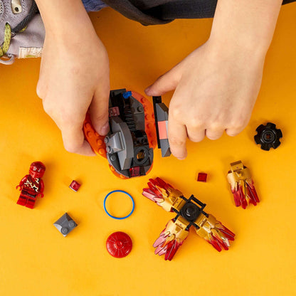 Child assembling LEGO® NINJAGO Spinjitzu Burst Kai Spinner Toy 70686 with colorful pieces and accessories on a yellow background.