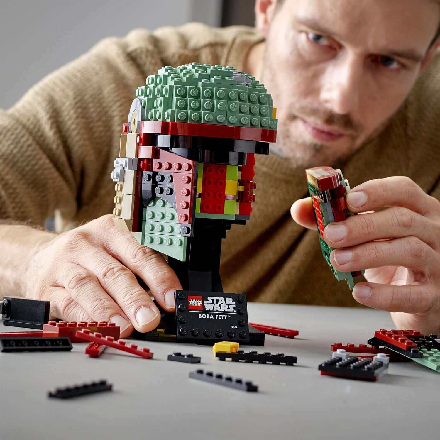 Adult builder assembling LEGO® Star Wars Boba Fett Helmet 75277 with colorful bricks and pieces, showcasing intricate details.
