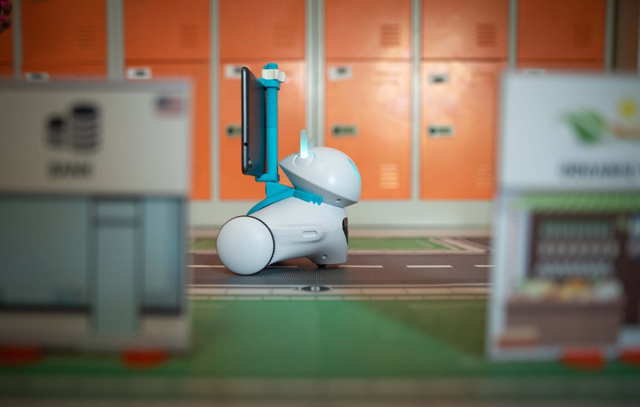 A small robot with a monitor navigating a miniature city street with orange lockers in the background.