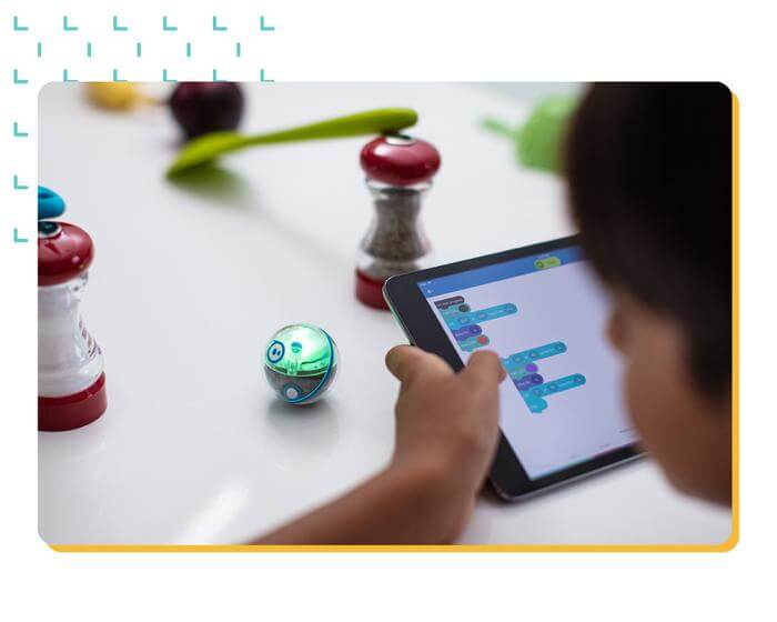 Child using a tablet to control a Sphero Mini robotic ball with coding blocks, surrounded by kitchen objects.
