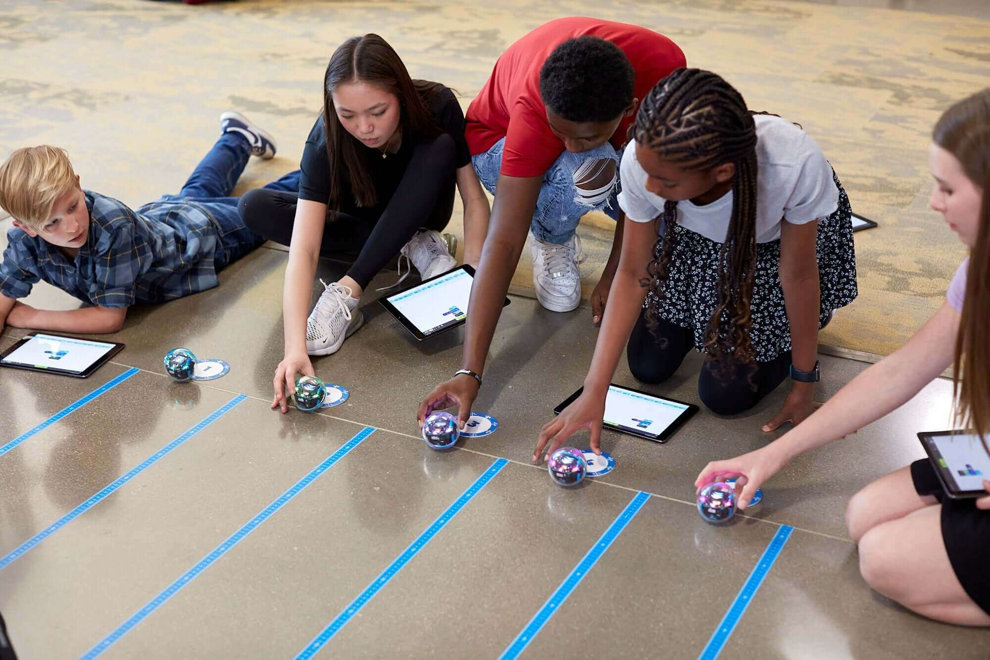 Students engaging in hands-on coding activities with Sphero robots on the floor using tablets.