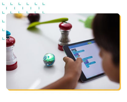 Child using a tablet to control a Sphero Mini robotic ball with coding blocks, surrounded by kitchen objects.