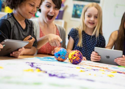 Excited children and teacher engaging in creative activity with tablets and colorful art projects.