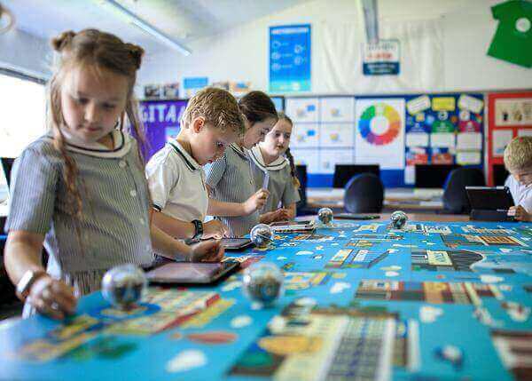 Children engaging with technology in a classroom, using tablets and interactive learning tools on a colorful table.