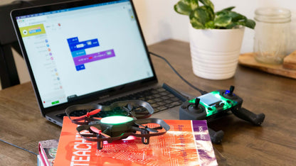 A laptop displaying coding software next to a drone and controller on a wooden table, with educational books underneath.